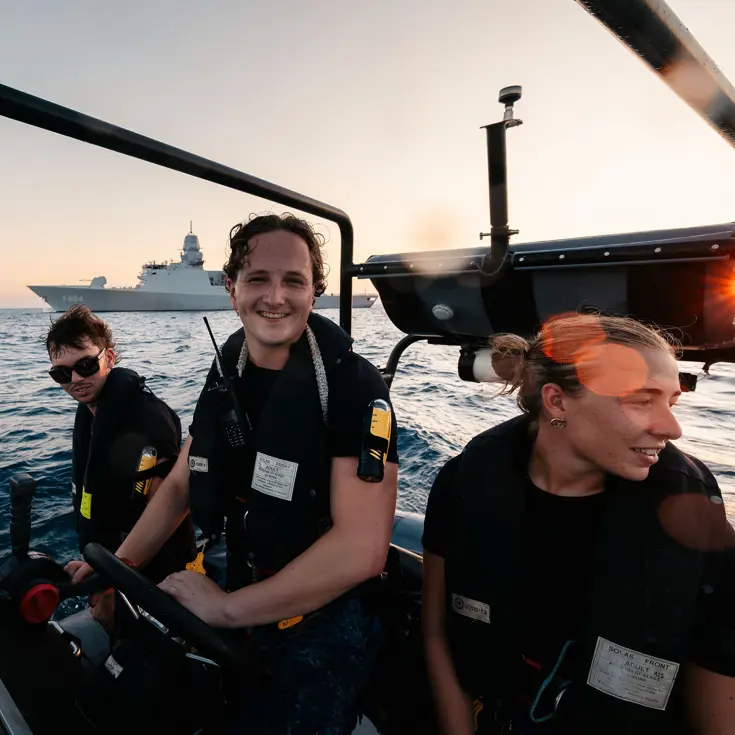 3 marinemensen varen met een RHIB, op de achtergrond ligt Zr.Ms. De Ruyter op zee.