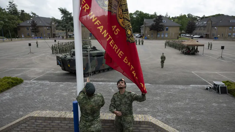 Twee militairen in camouflage hijsen een rode vlag met gouden tekst en embleem op een plein met militaire voertuigen en een opgestelde groep soldaten.