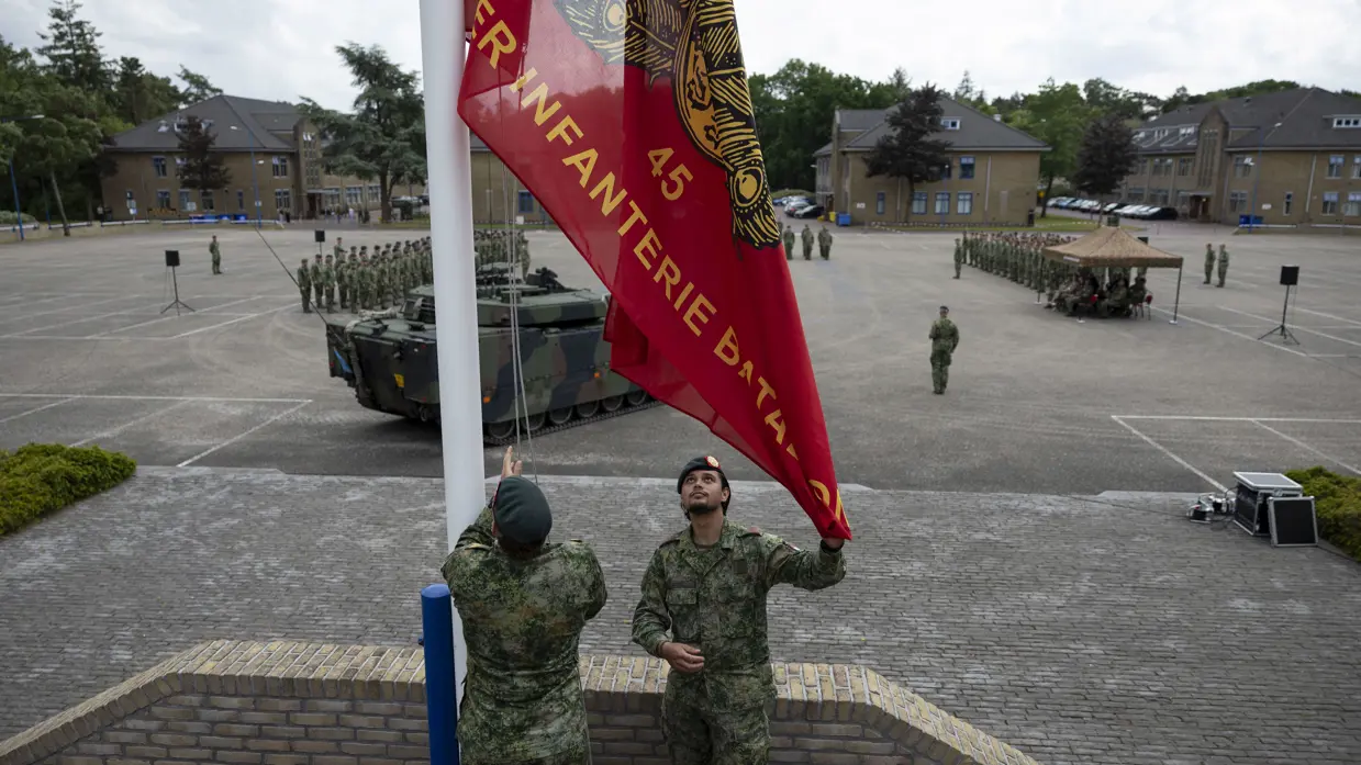 Twee militairen in camouflage hijsen een rode vlag met gouden tekst en embleem op een plein met militaire voertuigen en een opgestelde groep soldaten.
