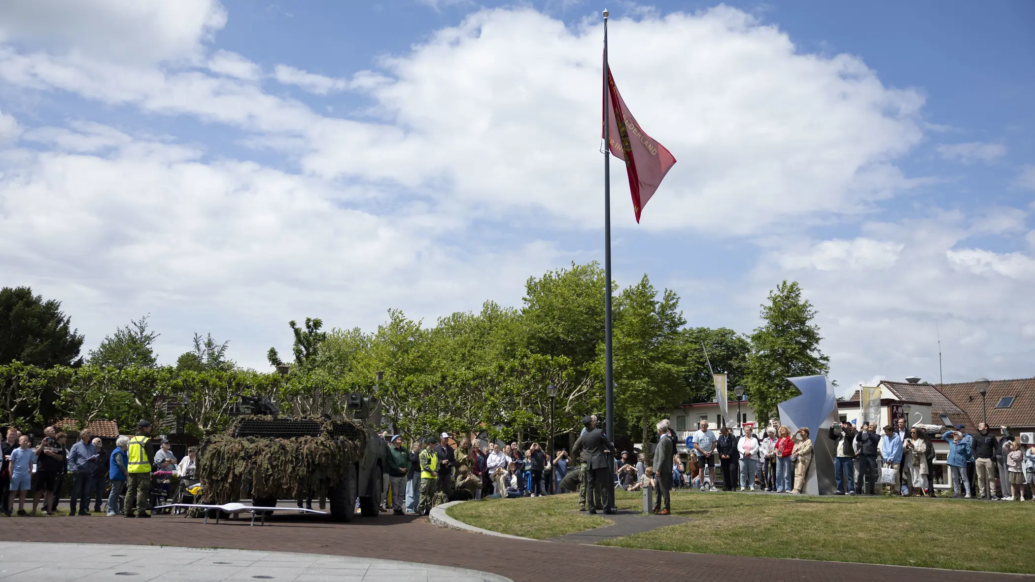 Een groep mensen kijkt naar een gecamoufleerde pantserwagen met een rode vlag op een zonnig plein met bomen en huizen.