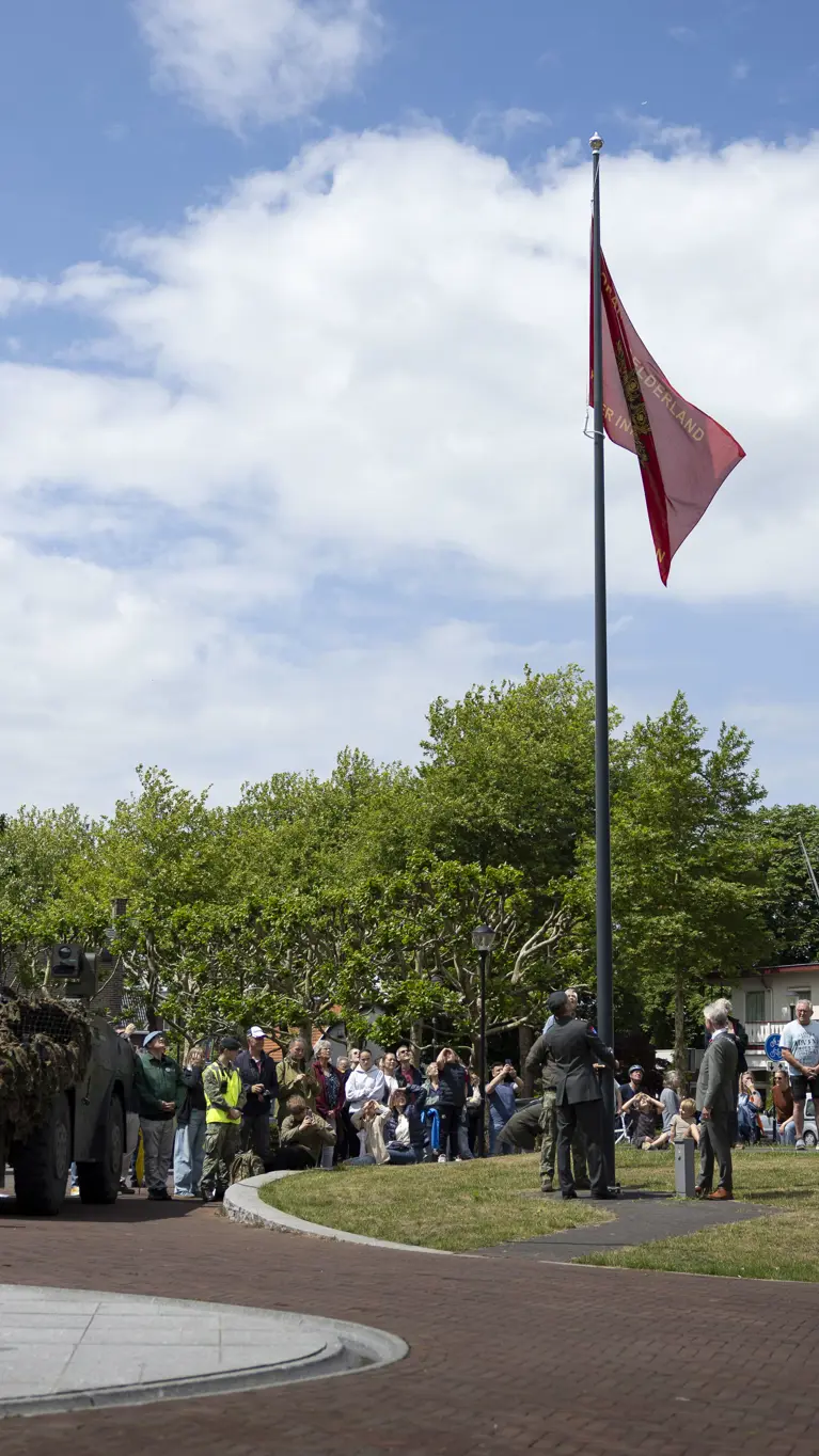 Een groep mensen kijkt naar een gecamoufleerde pantserwagen met een rode vlag op een zonnig plein met bomen en huizen.