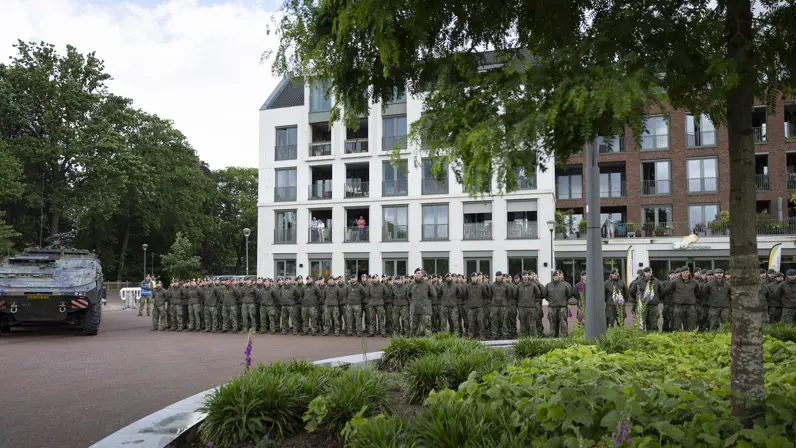 Een rij soldaten in uniform staat in de houding voor een wooncomplex, met een gepantserd voertuig en groen struikgewas.