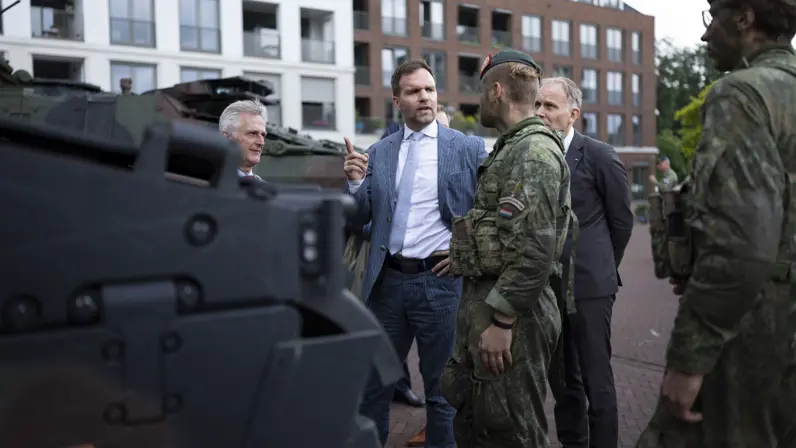 Een man in pak praat met soldaten in camouflage bij een gepantserd voertuig, met woonblokken op de achtergrond.