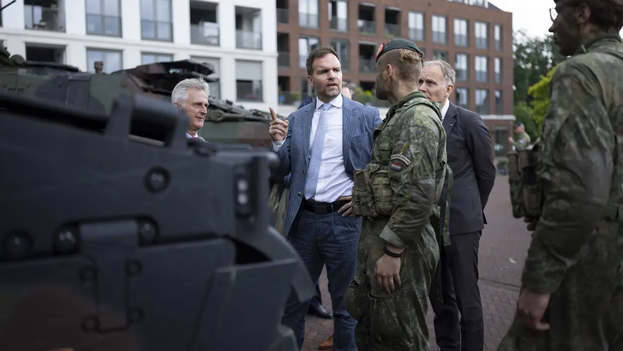 Een man in pak praat met soldaten in camouflage bij een gepantserd voertuig, met woonblokken op de achtergrond.