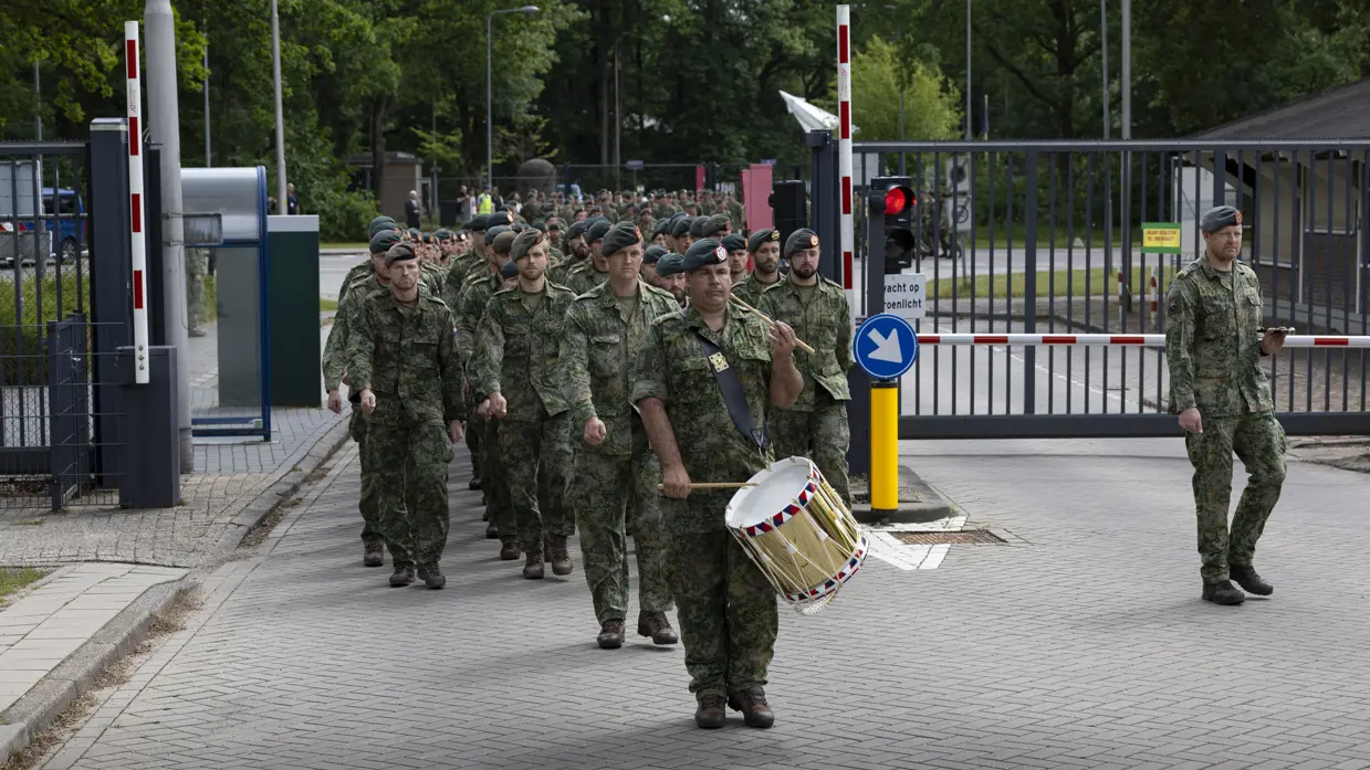 Soldaten in camouflage lopen in formatie door een hek, een drummer met trommel vooraan, bomen en gebouwen op de achtergrond. 