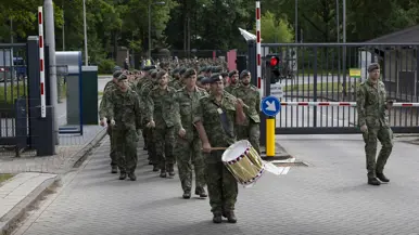 Soldaten in camouflage lopen in formatie door een hek, een drummer met trommel vooraan, bomen en gebouwen op de achtergrond. 