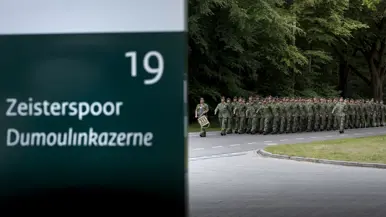 Soldaten in camouflage lopen in formatie langs een weg, met een bord "Zeisterspoor Dumoulinkazerne" en groen bos.