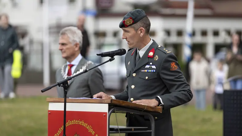 Een man in militair uniform met groene baret houdt een toespraak achter een lessenaar met microfoon, een andere man met ketting kijkt toe.