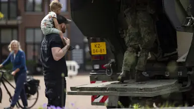 Een man draagt een kind op zijn schouders bij een militaire vrachtwagen, terwijl een soldaat instapt. Op de achtergrond fietst een vrouw.