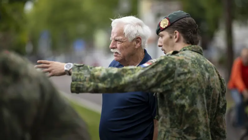 Een oudere man in donkerblauw overhemd en een jongere man in camouflageuniform met groene baret praten buiten, de oudere wijst naar links.