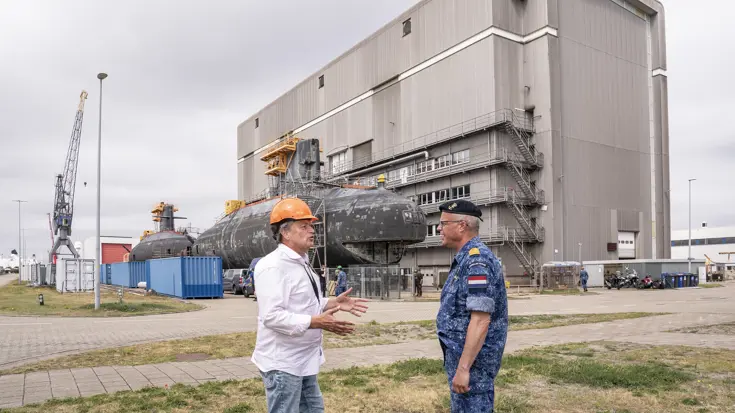 Een burger met oranje bouwhelm op is in gesprek met een marineman, voor een oude onderzeeboot.