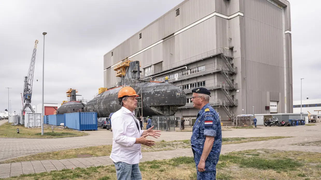 Een burger met oranje bouwhelm op is in gesprek met een marineman, voor een oude onderzeeboot.