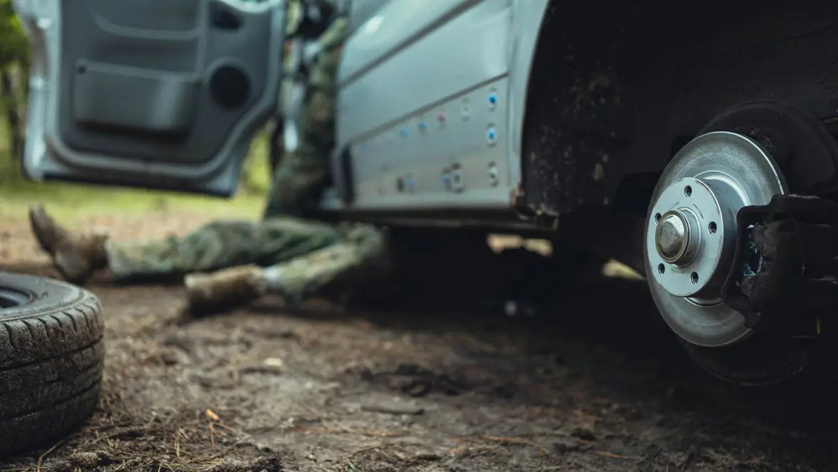 Een militair ligt onder een grijze bus in het bos om deze te repareren.