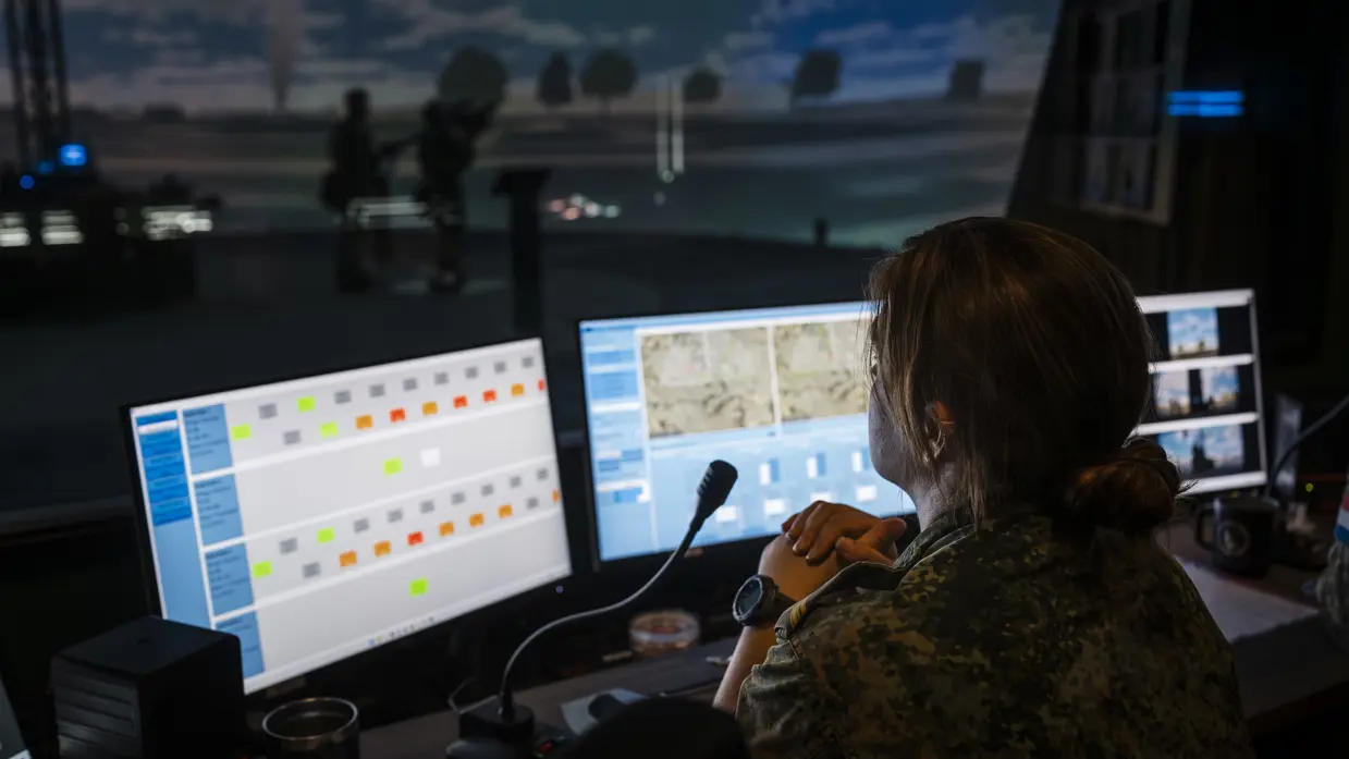 Een vrouwelijke militair kijkt over haar schermen naar de ruimte waarin de Stingerschutters rondlopen.