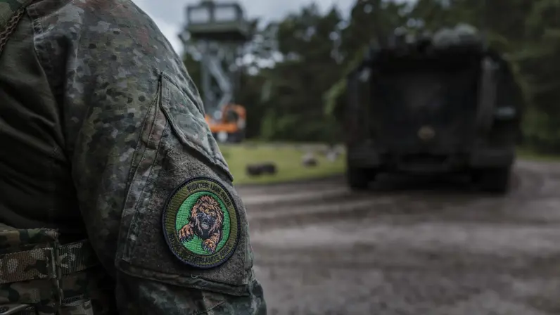 Close-up van de oefenbadge van Fighter Lion 2026 op de mouw van een militair. Op de achtergrond staat een militair voertuig op het oefenterrein.