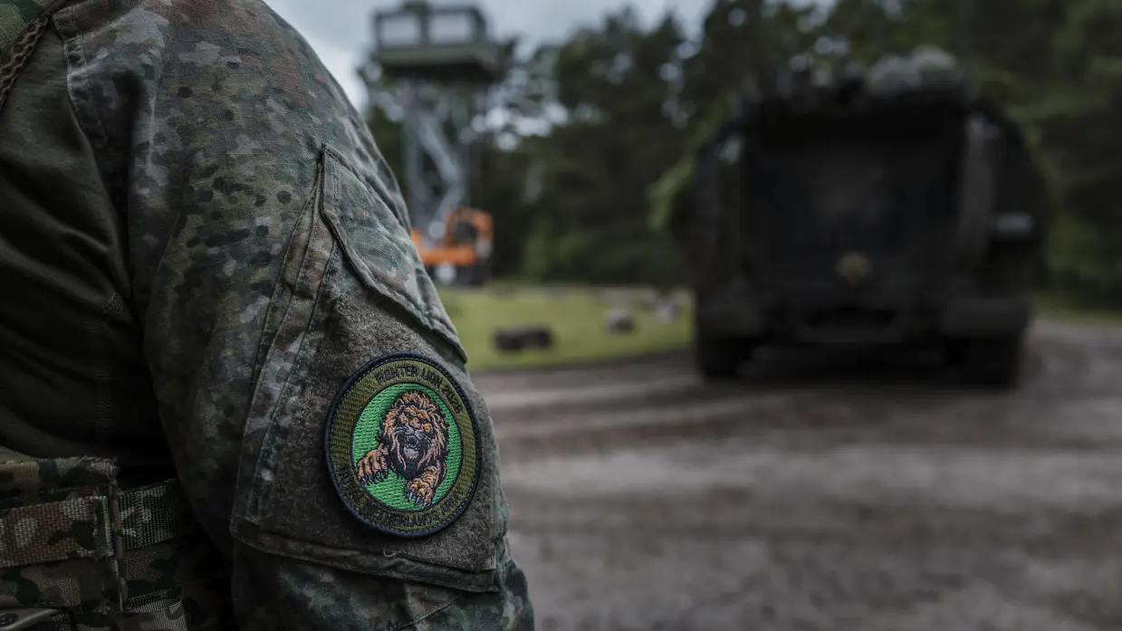Close-up van de oefenbadge van Fighter Lion 2026 op de mouw van een militair. Op de achtergrond staat een militair voertuig op het oefenterrein.