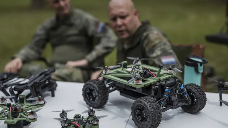 Een onbemand voertuig met een geïntegreerde drone staat op tafel tijdens een demonstratie van TechDevOps, terwijl kapitein Max en luitenant-kolonel Roeland op de achtergrond uitleg geven.