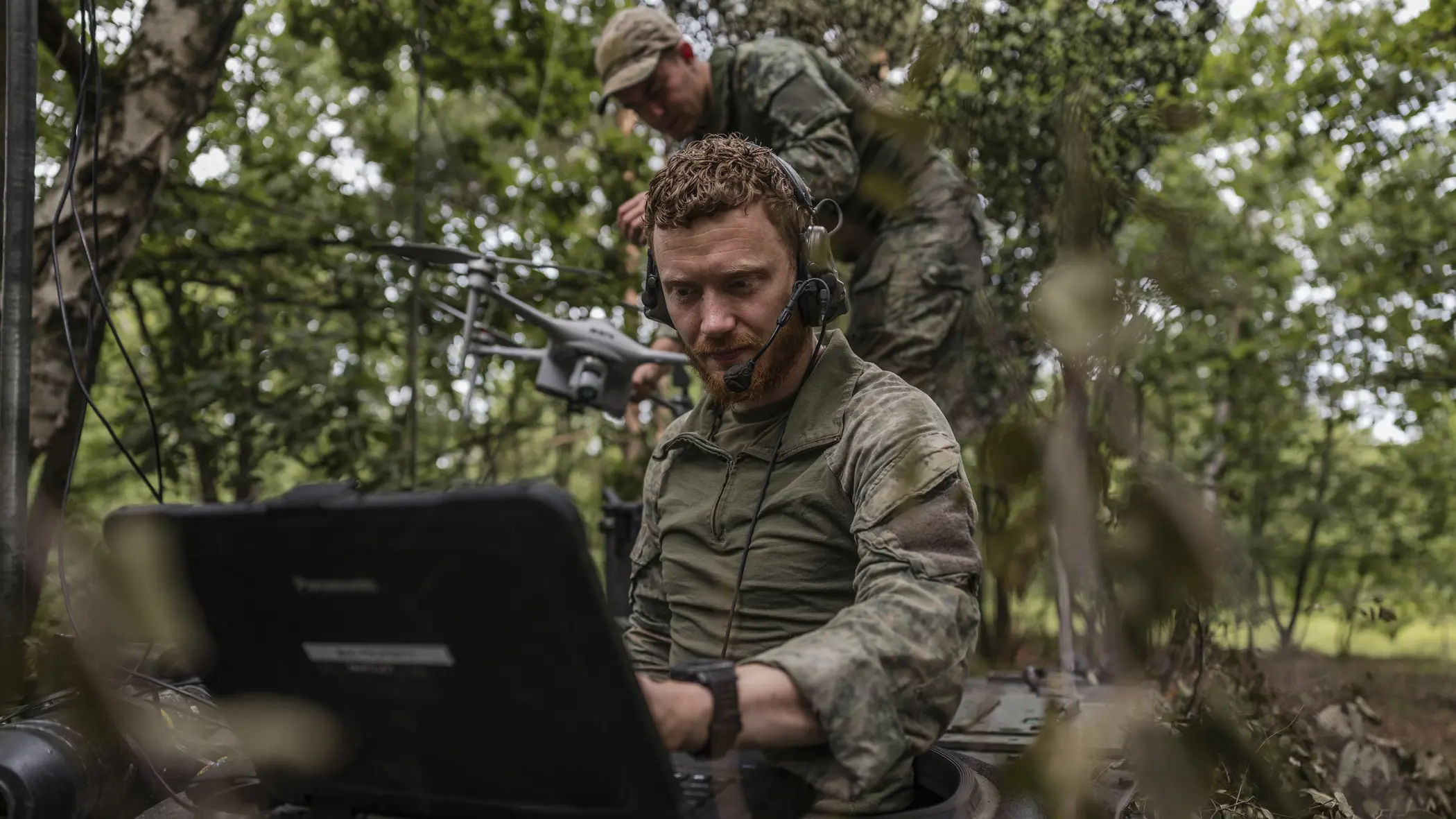  Een Forward Observer werkt vanaf een gecamoufleerde positie met een robuuste laptop, terwijl op de achtergrond een collega een verkenningsdrone gereedmaakt voor een vlucht tijdens een oefening.