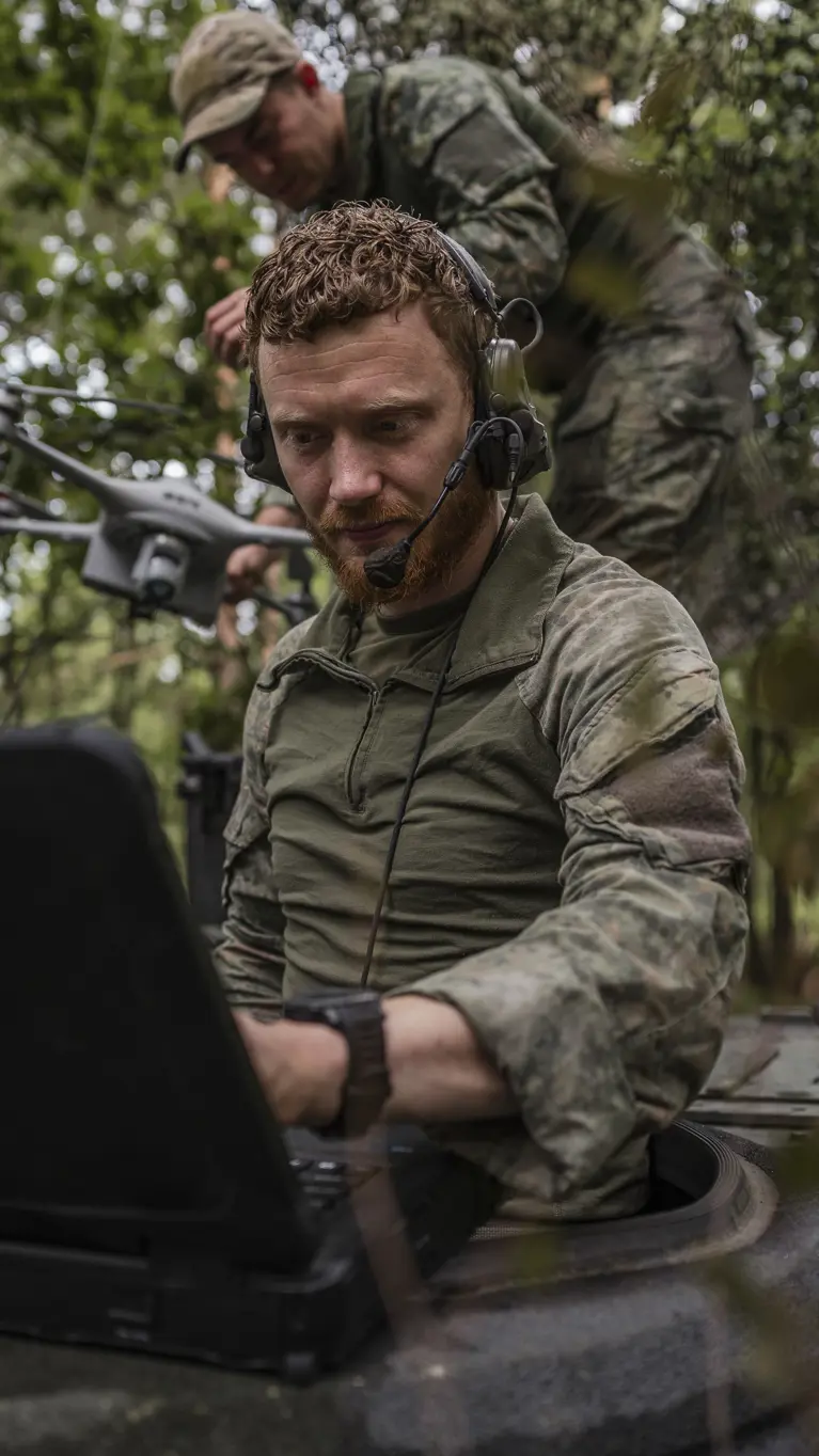  Een Forward Observer werkt vanaf een gecamoufleerde positie met een robuuste laptop, terwijl op de achtergrond een collega een verkenningsdrone gereedmaakt voor een vlucht tijdens een oefening.
