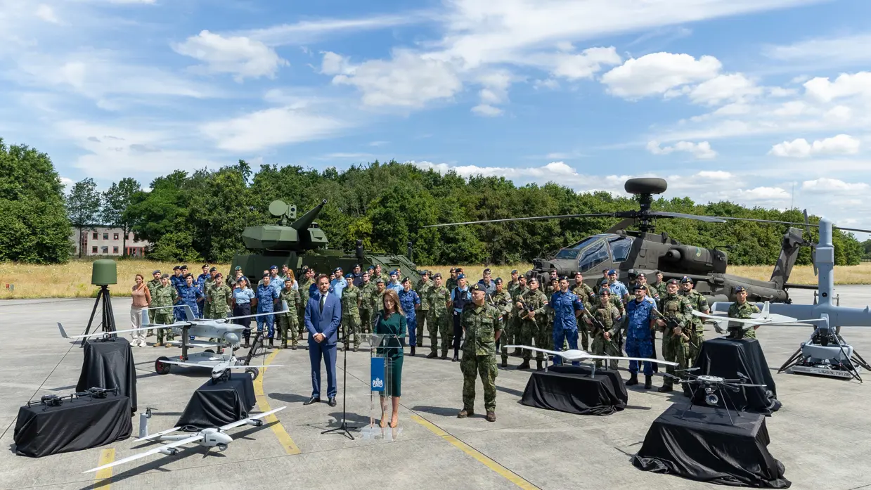 De minister presenteert de Defensienota met naast haar de stattssecretaris en de Commandant der Strijdkrachten. Achter hen staat een brede delegatie militairen en een Apache-gevechtshelikopter.
