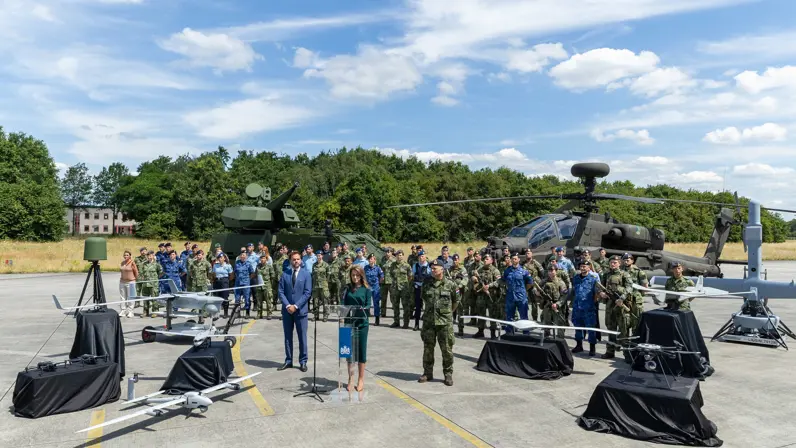 De minister presenteert de Defensienota met naast haar de stattssecretaris en de Commandant der Strijdkrachten. Achter hen staat een brede delegatie militairen en een Apache-gevechtshelikopter.