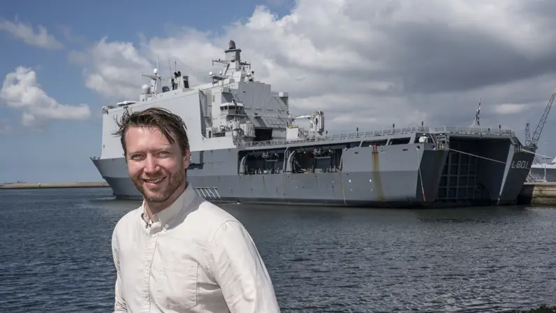 Man in witte blouse staat op kade marinebasis. Achter hem ligt een groot marineschip.