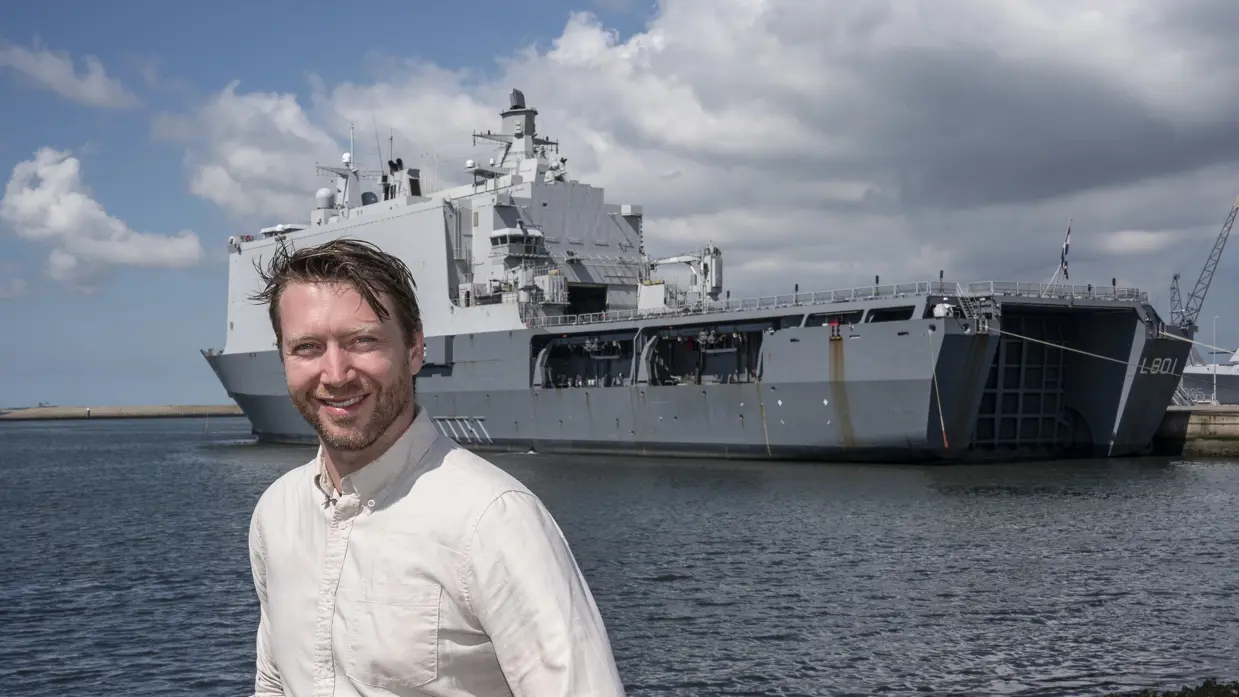 Man in witte blouse staat op kade marinebasis. Achter hem ligt een groot marineschip.