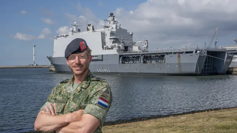 Marinier met baret op de kade met handen over elkaar. Op de achtergrond een groot marineschip.