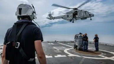Een marinecollega kijkt naar een helikopter die boven een scheepsdek hangt.