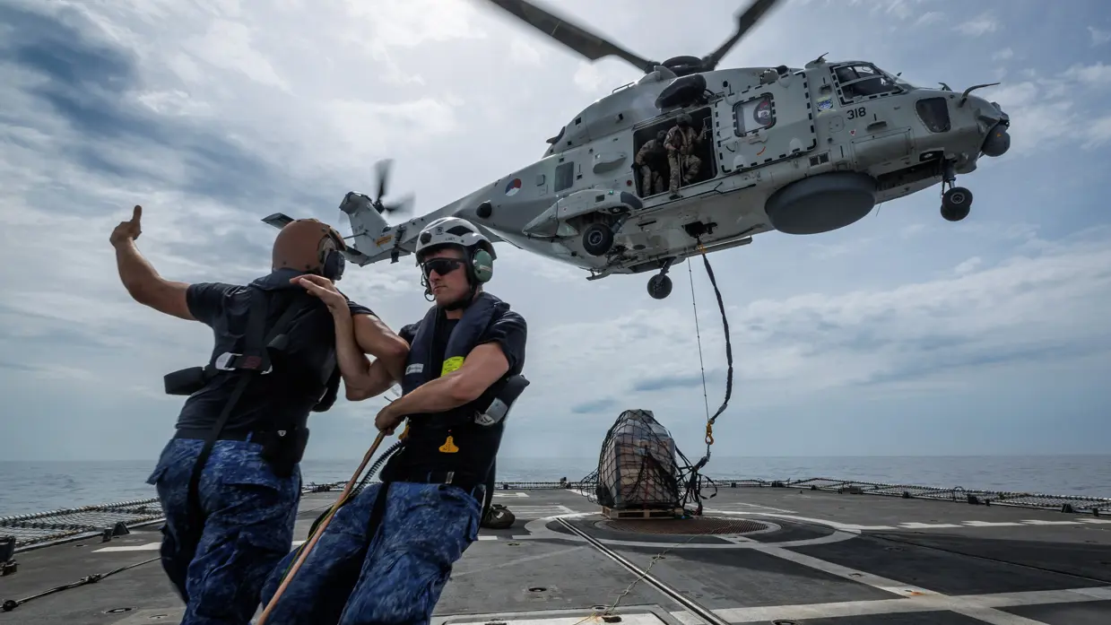 Een helikopter hangt in de lucht boven een pallet met goederen. 2 marinemensen zetten zich schrap tegen de wind van de rotorbladen.