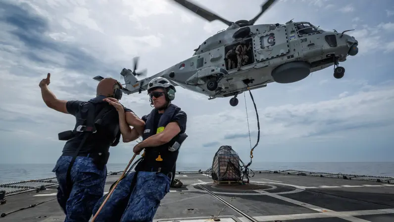 Een helikopter hangt in de lucht boven een pallet met goederen. 2 marinemensen zetten zich schrap tegen de wind van de rotorbladen.