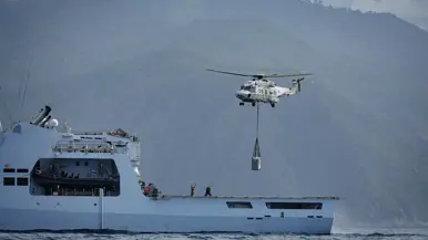 Er hangt een helikopter boven het achterdek en tilt een vracht de lucht in. 
