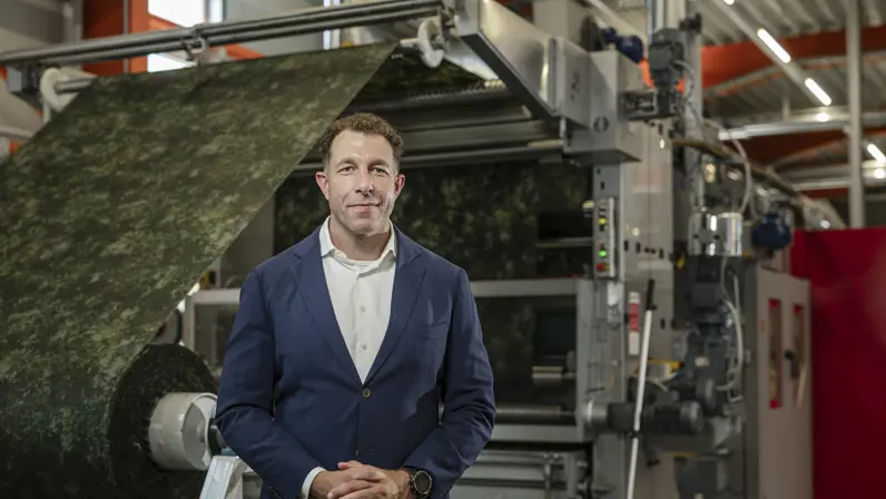Man met blauw colbertjas poseert in fabriekshal. Op de achtergrond wordt een grote rol stof in camouflagepatroon door een machine geleid.