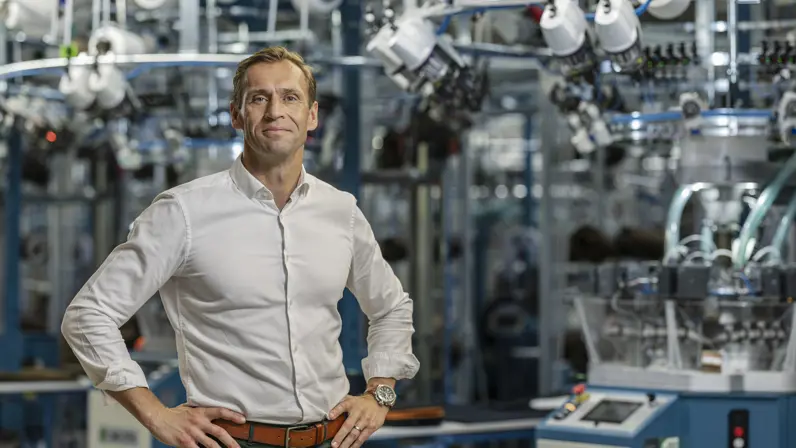 Man in witte blouse poseert met handen in de zij in fabriekshal, Op de achtergrond staan grote machines.