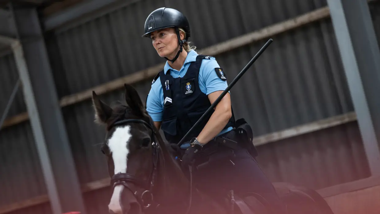 Een vrouw met een blauw marechausseeshirt aan rijdt op een zwart paard. Haar uitgestoken wapenstok raakt een staander met kussens erop.