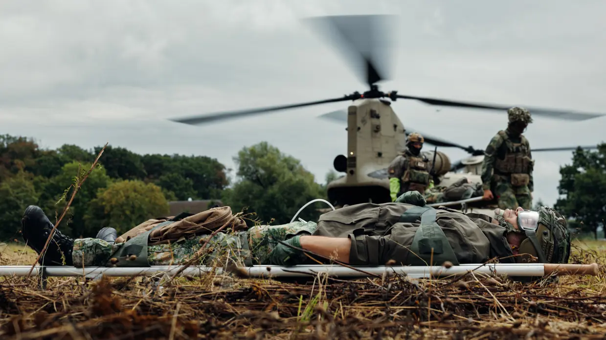 Een militair ligt op een brancard terwijl twee militairen in de achtergrond bij een helikopter zich klaarmaken.