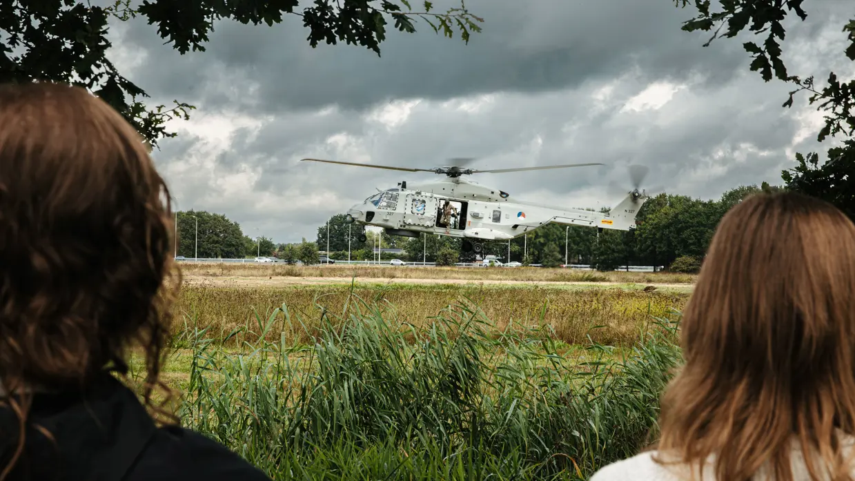 Twee personen in de voorgrond kijken richting een gelande helikopter.
