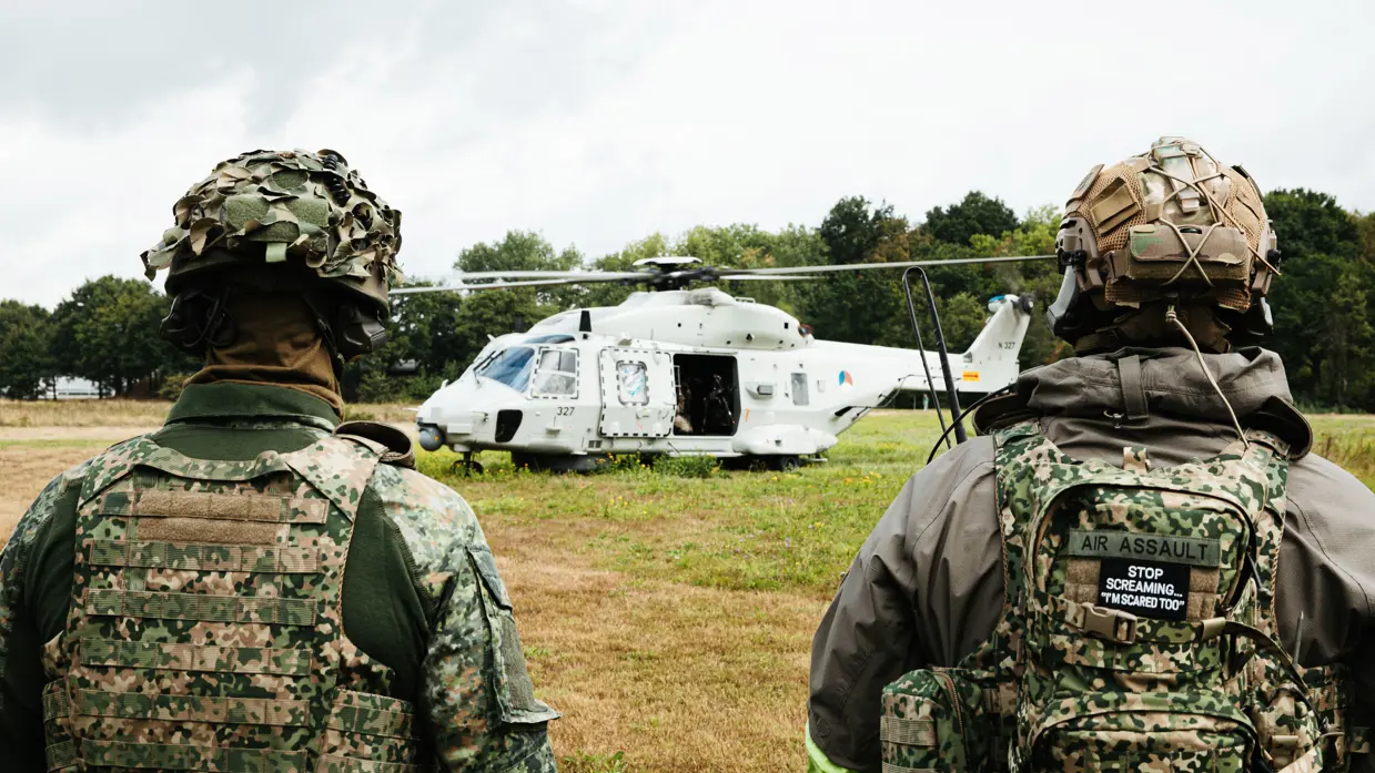 Twee militairen op de voorgrond kijken richting een gelande helikopter.
