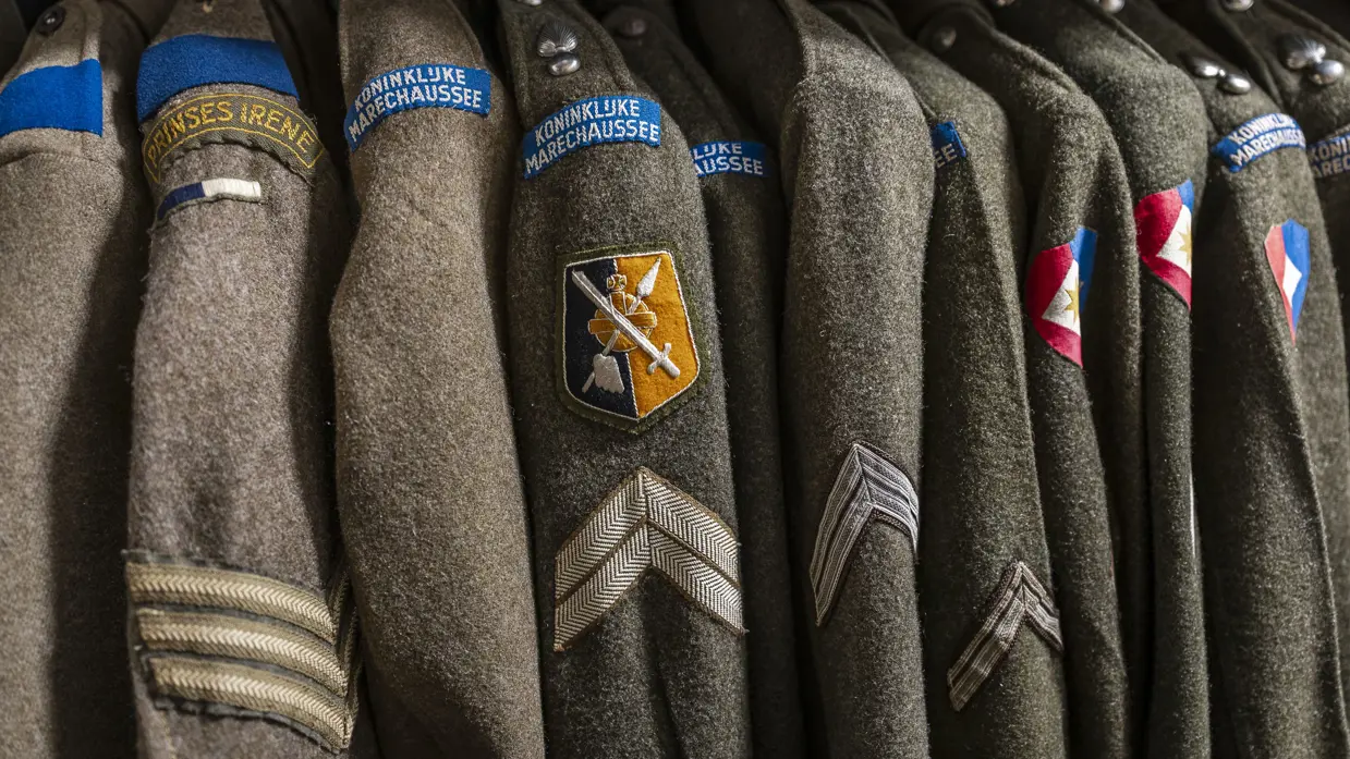 Close-up van een rij groene, wollen uniformen met een badge van de Koninklijke Marechaussee.