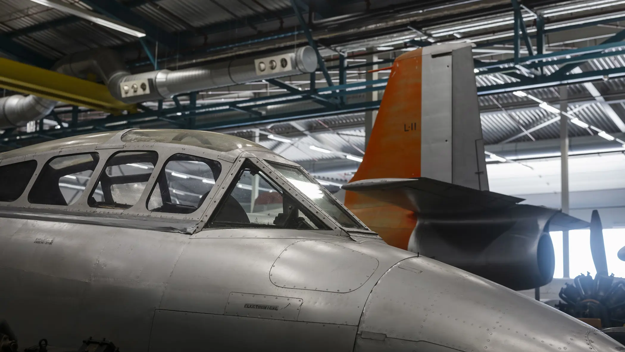 Close-up van een cockpit van een oud jachtvliegtuig met daarachter het staartstuk van een ander toestel.