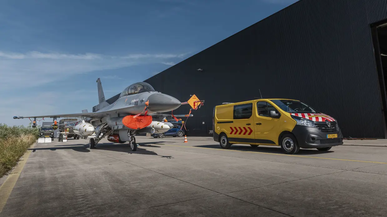 Een F-16 staat naast het depot, naast het toestel staat een geel bestelbusje.