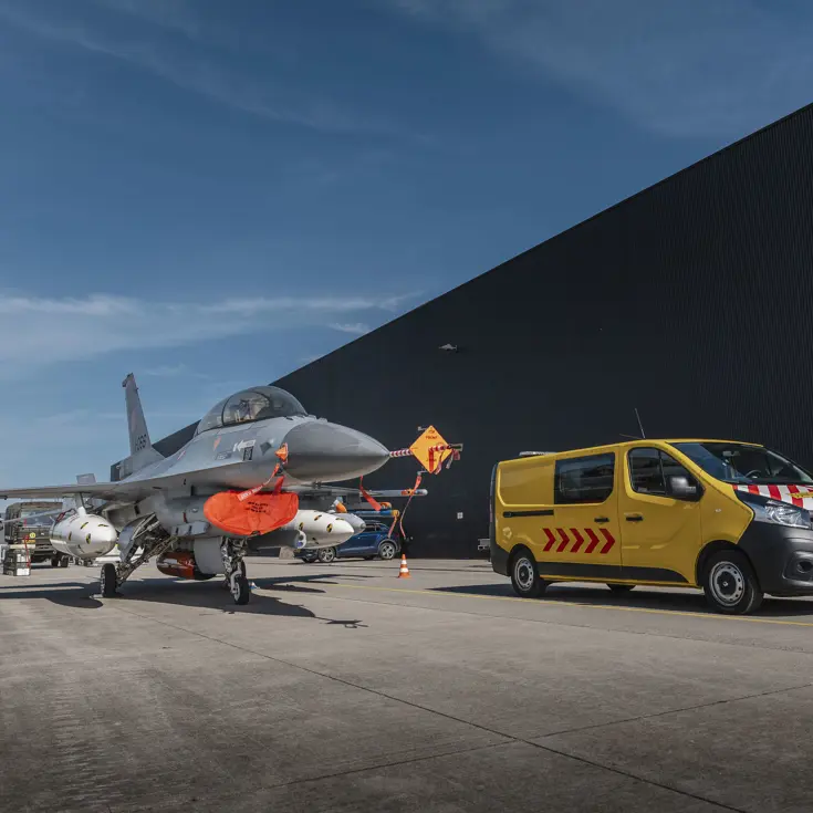Een F-16 staat naast het depot, naast het toestel staat een geel bestelbusje.