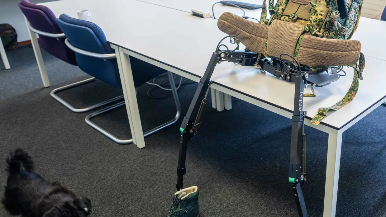 Links: een exoskelet met camouflage rugzak en groene bergschoenen staat op een tafel in een klaslokaal met stoelen en tafels.    Rechts een groene bergschoen met een zwarte metalen meetlat ernaast staat op een grijze vloer, tweede schoen op de achtergrond. 