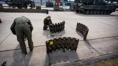 Twee soldaten in groene overall monteren een zware, metalen structuur op een betonnen oppervlak met tanks op de achtergrond.
