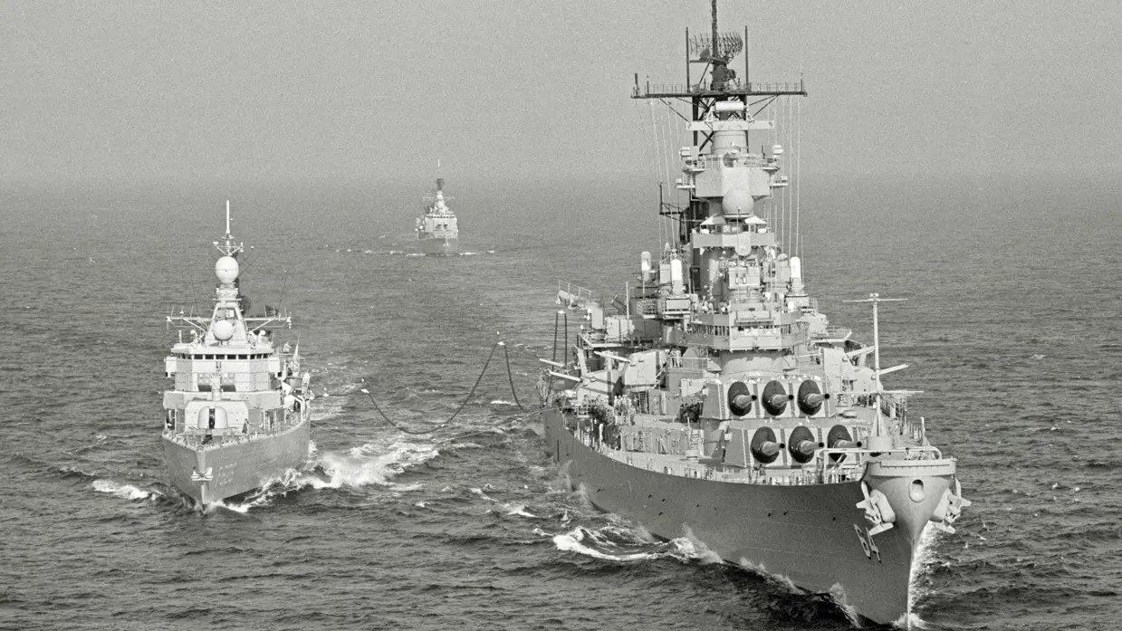  Drie oorlogsschepen varen op ruwe zee: twee grote schepen voorop, verbonden door kabels, en een kleiner schip op de achtergrond.