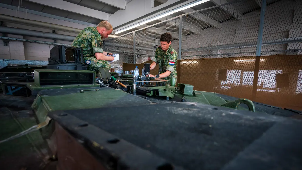 Twee cavaleristen in camouflage-uniform werken aan apparatuur op een groen pantservoertuig in een verlichte loods.