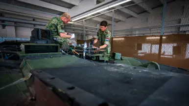 Twee cavaleristen in camouflage-uniform werken aan apparatuur op een groen pantservoertuig in een verlichte loods.