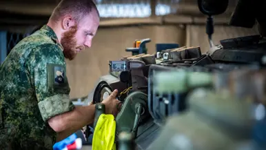 Een militair in camouflage-uniform werkt geconcentreerd aan een mechanisch onderdeel in een werkplaats met gereedschap.
