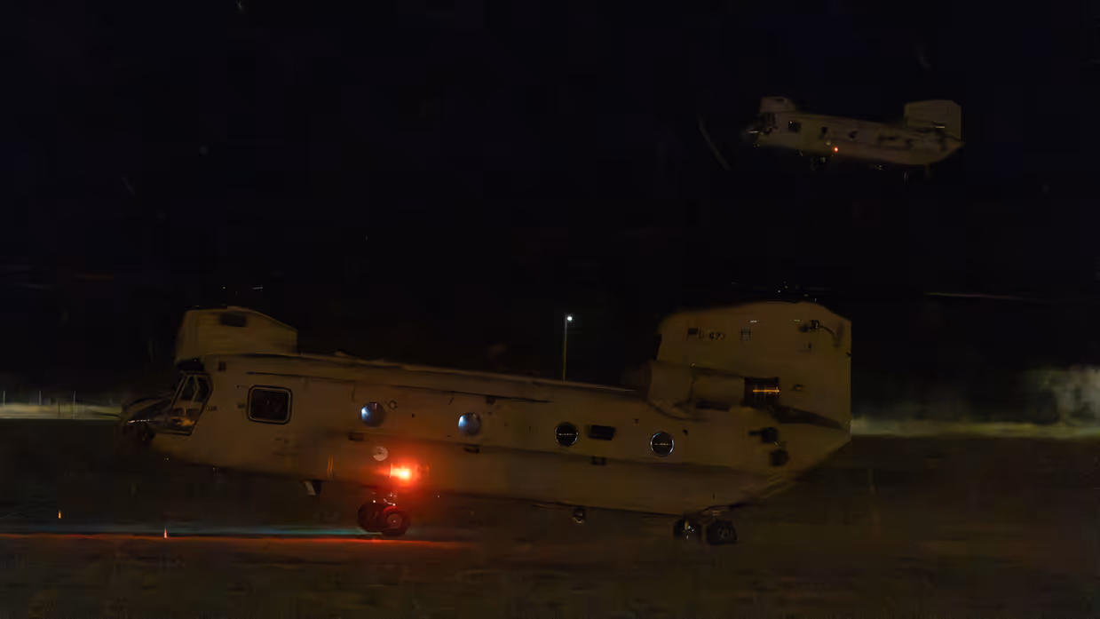 Twee Chinooks in het duister. Een in de lucht, de ander komt net los van het grasveld.