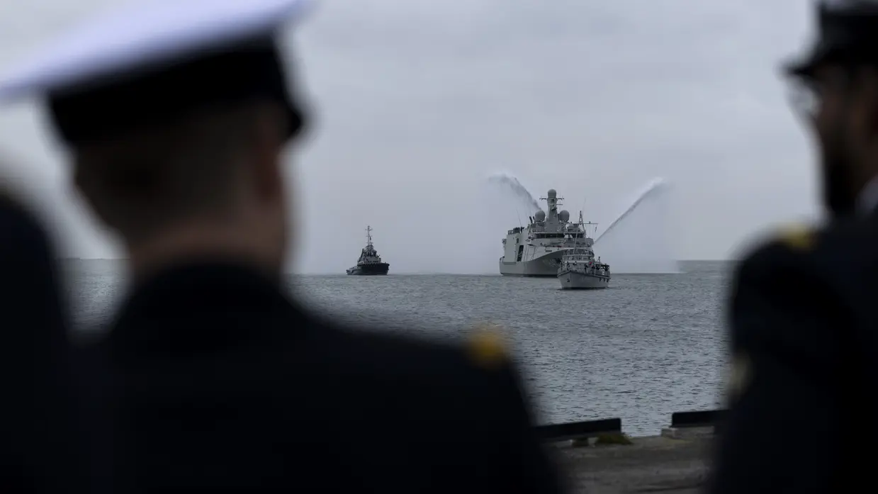 De Vlissingen met daarnaast een sleepboot, gezien vanaf de kade. Daarnaast het grijze marineschip van dichterbij, de sleepboot er nog altijd naast varend.